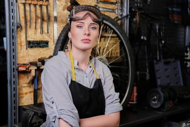 Zdjęcie portret pięknej kobiety brunetka ubrany w ubranie robocze, fartuch i gogle, stojąc w warsztacie przed narzędziami ściennymi.