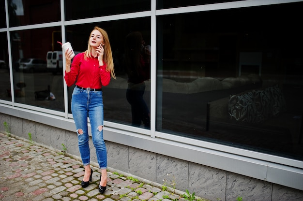 Portret pięknej kobiety w czerwonej bluzce i dżinsach rozmawia przez telefon komórkowy i trzyma filiżankę kawy poza ogromnym centrum handlowym.