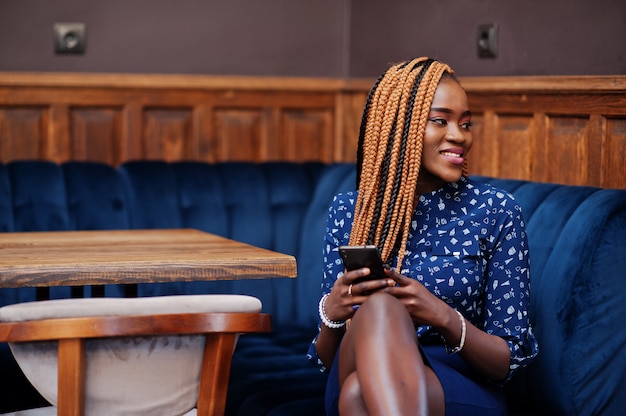 Portret pięknej młodej afrykańskiej kobiety biznesu z dredami, nosić na niebieską bluzkę i spódnicę, siedząc w kawiarni z telefonem komórkowym.