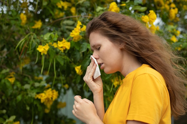 Portret pięknej młodej alergicznej kobiety cierpi na alergię na pyłki lub przeziębienie na tle drzewa kwitnącego naturalnego kwiatu wiosną lub słonecznym letnim dniem kicha dmuchanie katarem przeciera oczy