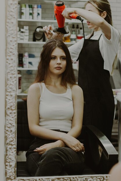 Portret pięknej młodej brunetki siedzącej na krześle przed lustrem, na którym fryzjerka suszy włosy suszarką z grzbietem, robiąc ślubną fryzurę z bliska.