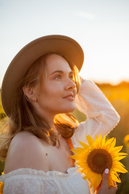 Portret pięknej młodej kobiety 33 lata w kapeluszu na polu słonecznika o zachodzie słońca Szczęśliwy model w białej sukni na letni wieczór w przyrodzie Ciepły