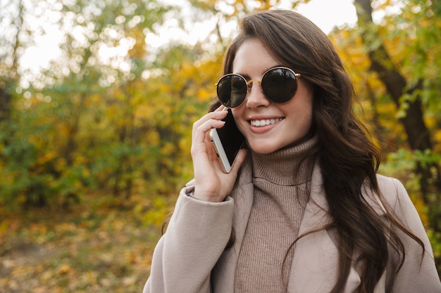 Portret pięknej młodej kobiety na zewnątrz w zielonym parku noszenia okularów przeciwsłonecznych rozmawia przez telefon.