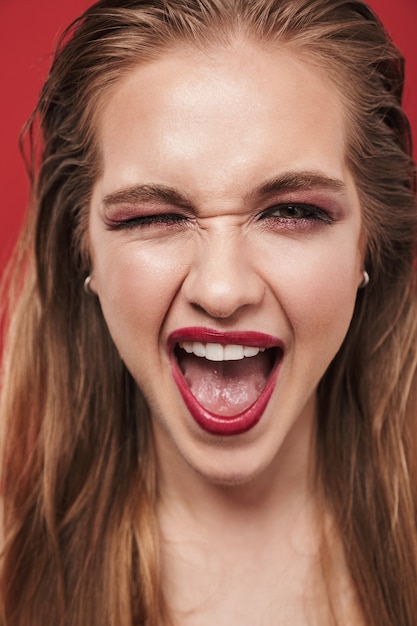 Portret pięknej młodej kobiety szczęśliwy emocjonalny roześmiany jasny makijaż czerwona szminka pozowanie na czerwonej ścianie mrugając.