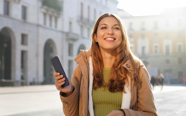 Portret pięknej młodej kobiety trzymającej smartfona na tle miasta, patrząc w bok i odwracając myślenie