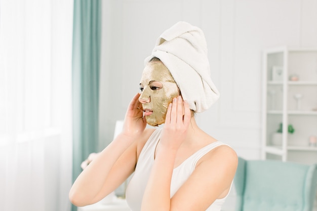 Portret pięknej młodej kobiety w biały ręcznik na głowie z nagim makijażu i kosmetycznych papieru złota maska na twarz.