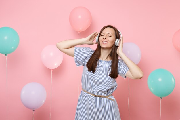 Portret pięknej młodej kobiety z zamkniętymi oczami ze słuchawkami w niebieskiej sukience słuchania muzyki na pastelowym różowym tle z kolorowych balonów. Urodziny wakacje party ludzie szczere emocje.