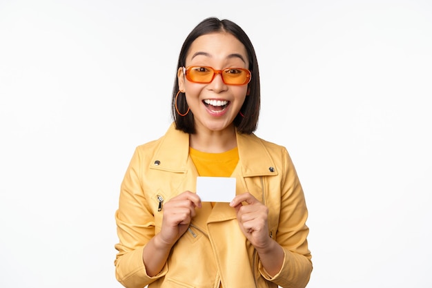 Portret pięknej nowoczesnej azjatykciej dziewczyny w okularach przeciwsłonecznych uśmiechający się szczęśliwy pokazujący kartę kredytową stojącą nad białym tłem kopii przestrzenią
