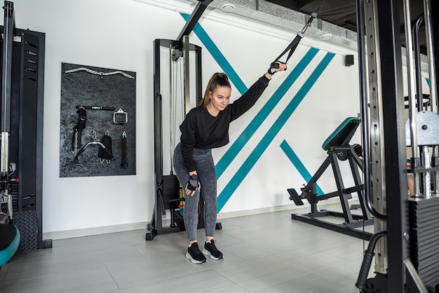 Portret pięknej pani fitness ćwiczącej na siłowni, wykonującej ćwiczenia, które pompują mięśnie pleców, uśmiecha się do sportu jak do życia