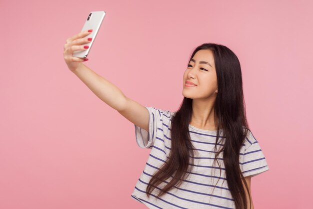 Portret pięknej pozytywnej azjatyckiej dziewczyny z długimi włosami brunetki i pięknym makijażem, biorąc selfie, uśmiechając się przyjaźnie, robiąc zdjęcie dla bloga o modzie. strzał w studio na białym tle na różowym tle