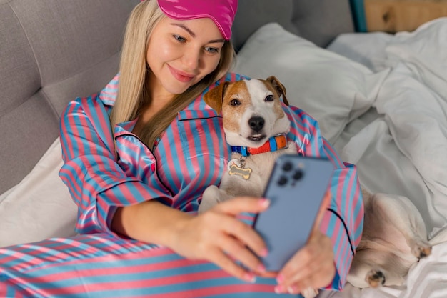 Portret pięknej radosnej kobiety w piżamie przytulającej psa za pomocą telefonu komórkowego do selfie uśmiechającego się leżącego na łóżku po śnie lub drzemce