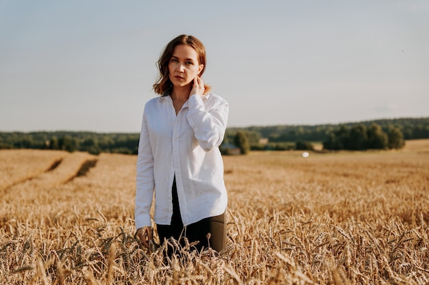 Portret pięknej rudowłosej dziewczyny w białej koszuli. Stoi na polu żyta w słoneczny dzień. Spokojny wyraz twarzy. Pojęcie ustępstw