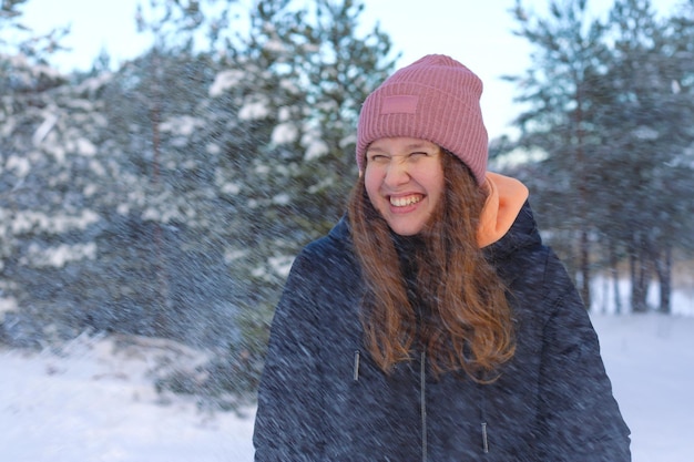 Portret pięknej, słodkiej dziewczyny w parku lub lesie na zewnątrz kobieta, która dobrze się bawi, śmiejąc się, uśmiechając się do nastolatka natury w ciepłym ubraniu Zamieć i spadające płatki śniegu