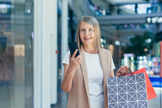 Portret Pięknej Starszej Kobiety Robiącej Zakupy W Centrum Handlowym Rozmawia Przez Telefon, Używa Telefonu, Trzyma W Rękach Papierowe Torby Na Zakupy Uśmiecha Się Zadowolona Z Zakupów