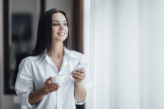 Portret Pięknej Szczęśliwej Dziewczyny Brunetka Rano Z Kawą I Telefonem W Pobliżu Okna.