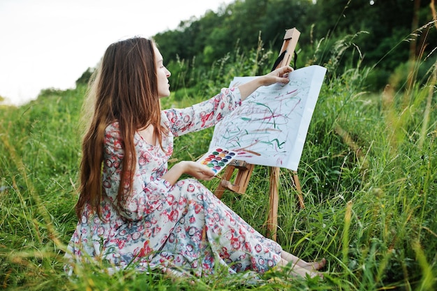 Portret pięknej szczęśliwej młodej kobiety w pięknej sukience siedzącej na trawie i malującej na papierze akwarelami