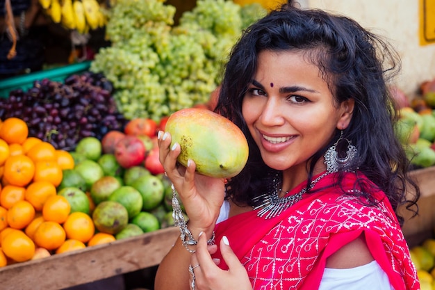 Portret pięknej tradycyjnej Indianki w czerwonej sukience sari wybierającej owoce mango na targu ulicznym w IndiachSzczęśliwa dziewczyna kupujący w supermarkecie sklep z owocamiegzotyczna wyprzedaż Kerala Goa