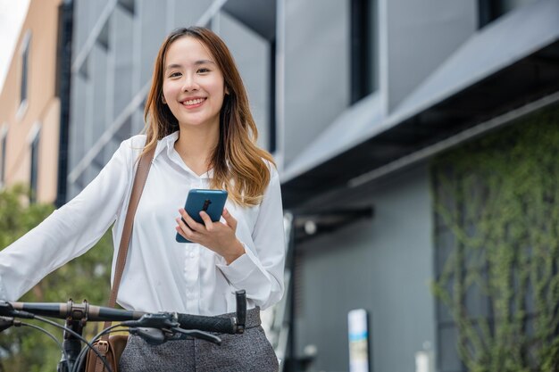 Portret pięknej uśmiechniętej bizneswoman dojeżdża na rowerze na zewnątrz za pomocą smartfona na rowerze miejskim idzie do pracy w biurze azjatycka bizneswoman stojąca na ulicy z rowerem trzymającym telefon komórkowy