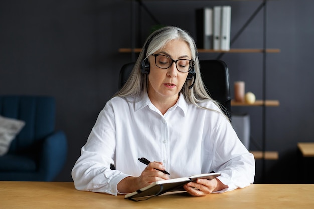 Portret pięknej uśmiechniętej starszej kobiety studiującej online. Edukacja online, praca zdalna, edukacja domowa. Siwowłosa starsza kobieta pisze w notesie. Osoby dorosłe studiują kursy online.