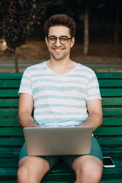 Portret Piękny Młody Mężczyzna Freelancer Patrząc Na Kamery, śmiejąc Się Podczas Pracy Na Swoim Laptopie Na świeżym Powietrzu W Parku Na ławce.