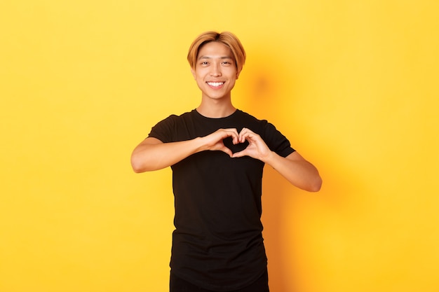 Portret piękny przystojny blond azjatycki mężczyzna, pokazując gest serca i uśmiechnięty, stojący żółtą ścianę