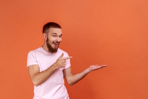 Portret podekscytowanego mężczyzny przedstawiającego obszar reklamowy na swojej dłoni i wskazującego miejsce kopiowania, trzymającego puste miejsce na reklamę, ubrany w różową koszulkę. Kryty studio strzał na białym tle na pomarańczowym tle.