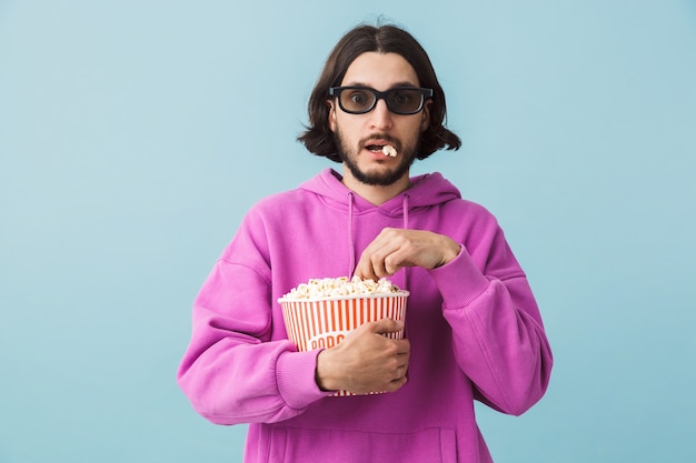 Portret podekscytowanego młodego brodatego mężczyzny w bluzie z kapturem, stojącego na białym tle nad niebieską ścianą, jedzącego popcorn, oglądającego film w okularach 3 d