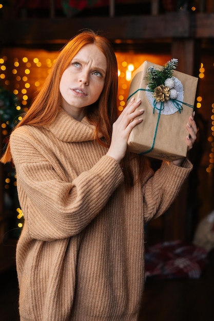 Portret podekscytowanej rudej młodej kobiety potrząsającej pudełkiem prezentów z zapakowanymi prezentami świątecznymi