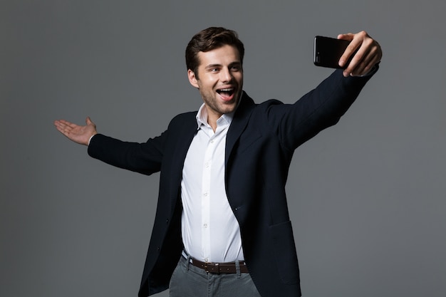 Portret podekscytowany młody biznesmen ubrany w garnitur na białym tle nad szarą ścianą, biorąc selfie