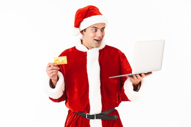 Portret podekscytowany młody człowiek ubrany w strój Świętego Mikołaja stojący na białym tle nad białą przestrzenią, trzymając laptopa