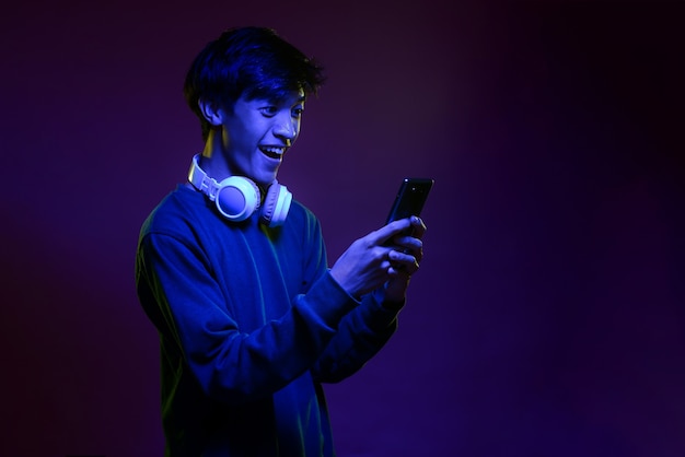 Portret podekscytowany przystojny młody azjatycki mężczyzna patrząc na telefon komórkowy noszenie słuchawek w świetle neonowym. Futurystyczne oświetlenie neonowe. Modny styl