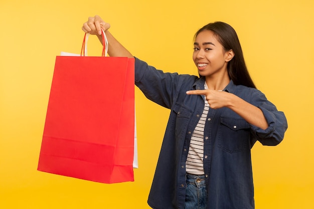 Portret podekscytowany, szczęśliwy shopper dziewczyna w stylowej dżinsowej koszuli wskazując torby, uśmiechając się do kamery, zadowolony z zakupów w sklepie z modą, sprzedaż oszczędności. strzał w studio na białym tle na żółtym tle