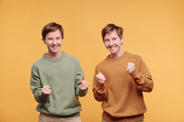 Portret podekscytowanych emocjonalnie braci bliźniaków w swobodnych swetrach wykonujących gesty tak na pomarańczowym tle