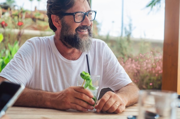 Portret pogodnego szczęśliwego dorosłego mężczyzny pijącego koktajl mojito w kawiarni podczas letnich wakacji spędzania wolnego czasu na świeżym powietrzu Turysta cieszący się relaksem Jeden dojrzały mężczyzna uśmiechający się siedzący przy stole