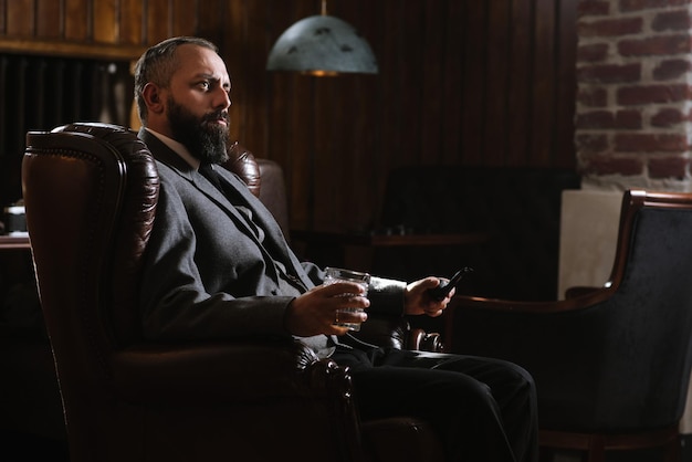 Portret poważnego brodatego mężczyzny z fajką trzymającego szklankę whisky w garniturze i siedzącego na dużym fotelu