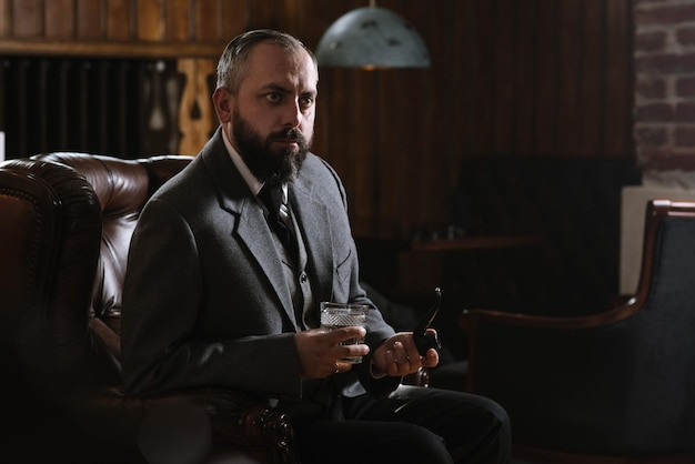 Portret poważnego brodatego mężczyzny z fajką trzymającego szklankę whisky w garniturze i siedzącego na dużym fotelu