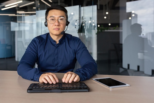 Portret poważnego, odnoszącego sukcesy specjalisty w biurze, azjatyckiego mężczyzny z telefonem słuchawkowym i klawiaturą