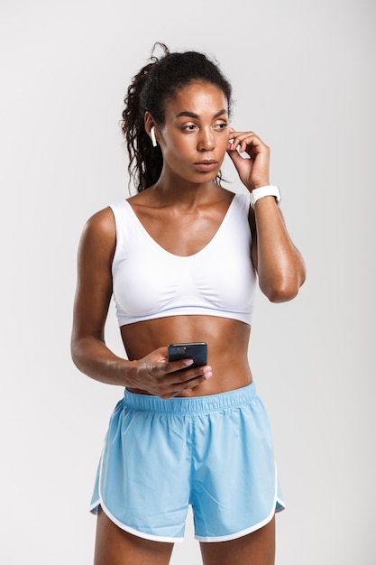Portret poważnej afroamerykańskiej kobiety w odzieży sportowej za pomocą telefonu komórkowego i słuchawek na białym tle nad białą ścianą