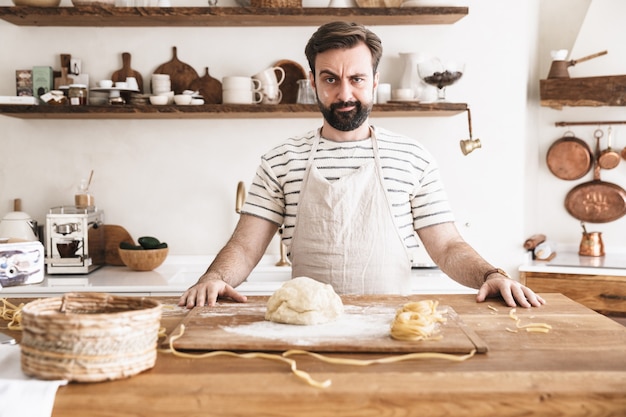 Portret Poważnej Brunetki Ubrany W Fartuch, Gotujący I Robiący Domowy Makaron Z Ciasta W Kuchni W Domu