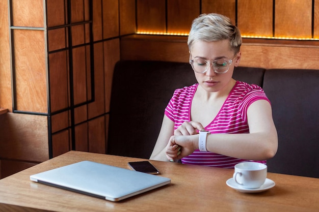 Portret poważnej młodej bussineswoman z krótkimi włosami w różowej koszulce siedzi w kawiarni sprawdzając czas na inteligentnym zegarku na nadgarstek i patrząc na kamerę z zdenerwowaną twarzą Kryty styl życia