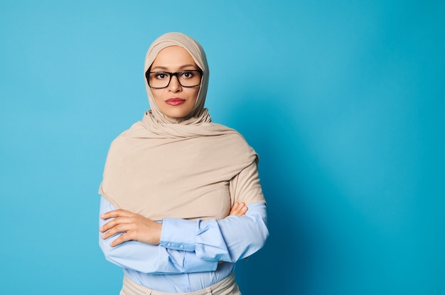 Portret poważnej młodej kobiety arabskiej noszenia hidżabu i okularach stojących ze skrzyżowanymi rękami na piersi