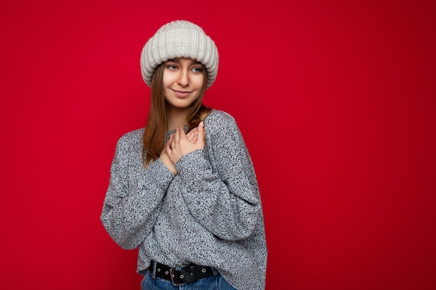 Portret pozytywne szczęśliwy młody piękny ciemny blond kobieta ze szczerymi emocjami na sobie szary dżersej i beżowy kapelusz na białym tle nad czerwonym tle z miejsca kopii i trzymając się za ręce na klatce piersiowej.