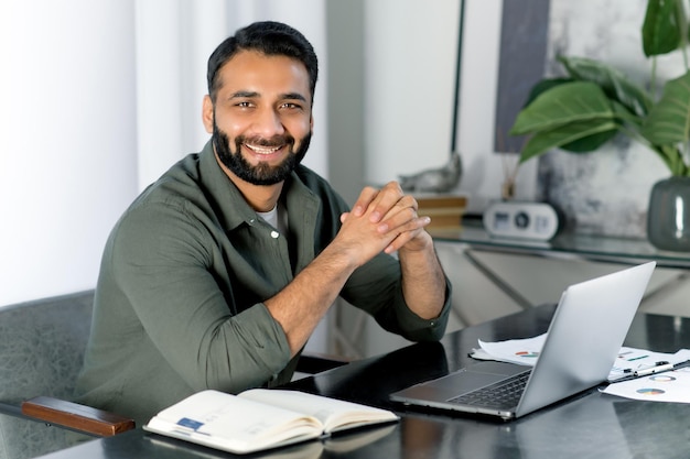 Portret pozytywnego, kreatywnego mężczyzny indyjskiego lub arabskiego, noszącego dorywczo stylowe ubrania, kierownik firmy, makler giełdowy, patrzy bezpośrednio w kamerę, uśmiechając się przyjazny, siedząc w nowoczesnym biurze