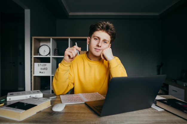 Portret pozytywnego młodzieńca w pomarańczowej bluzie siedzi przy laptopie i książki w sypialni z piórem w rękach pozuje do kamery Przystojny student chłopiec studiuje w domu i patrzy w kamerę