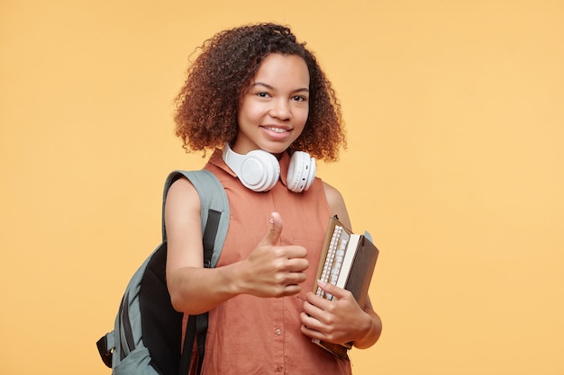 Portret Pozytywnej Afroamerykańskiej Dziewczyny Studentów Z Kręconymi Włosami, Trzymając Książki I Pokazując Kciuk W Górę Na żółtym Tle