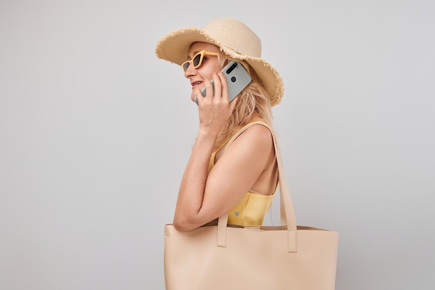 Portret pozytywnej dojrzałej blondynki w słomkowym kapeluszu żółtym topie i okularach przeciwsłonecznych trzymającej smartfona i torbę na zakupy odizolowaną na tle białego studia