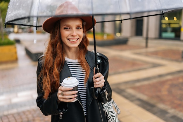 Zdjęcie portret pozytywnej imbirowej młodej kobiety w eleganckim kapeluszu trzymającej w ręku kubek z gorącą kawą stojącą z przezroczystym parasolem w zimnym jesiennym deszczu