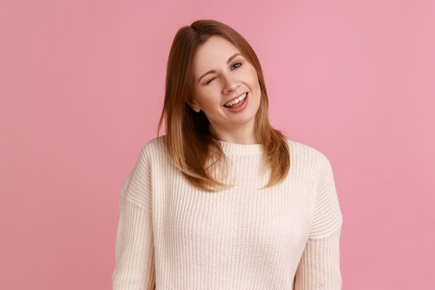 Portret pozytywnej optymistycznej blond kobiety stojącej i mrugającej żartobliwie, pozytywnej ekspresji, patrząc na kamerę, ubrana w biały sweter. Kryty studio strzał na białym tle na różowym tle.