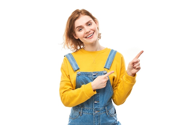 Portret pozytywnej rudej młodej kobiety w swobodnych dżinsach uśmiecha się wskazując palcem na pustą przestrzeń dla tekstu lub produktu na białym tle baner reklamowy