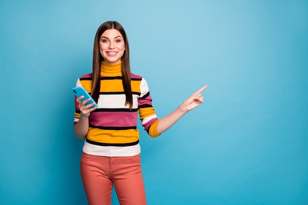 Portret pozytywnej wesołej dziewczyny wskazującej palcem wskazującym za pomocą smartfona polecam promocję w mediach społecznościowych online noś stylową modną odzież izolowaną na niebieskiej ścianie